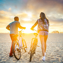 A man and woman walking their bikes on a beach at sunrise.